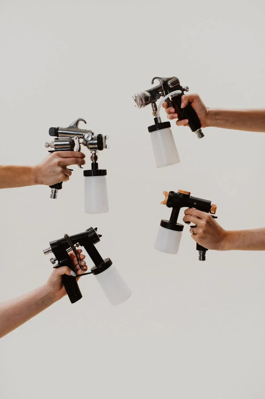 Four hands holding HVLP spray tan guns from below against a light background