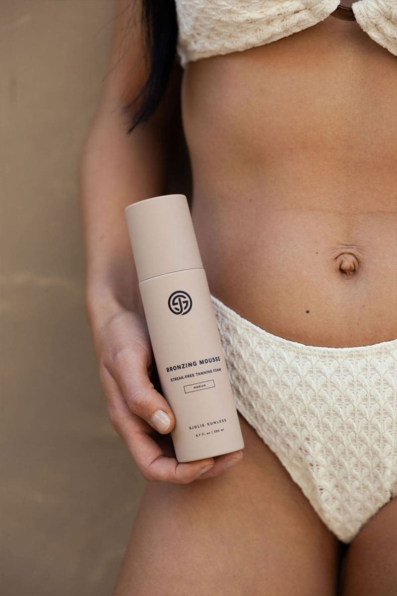 Model in a white bikini holding a pink bottle of SJOLIE Bronzing Mousse