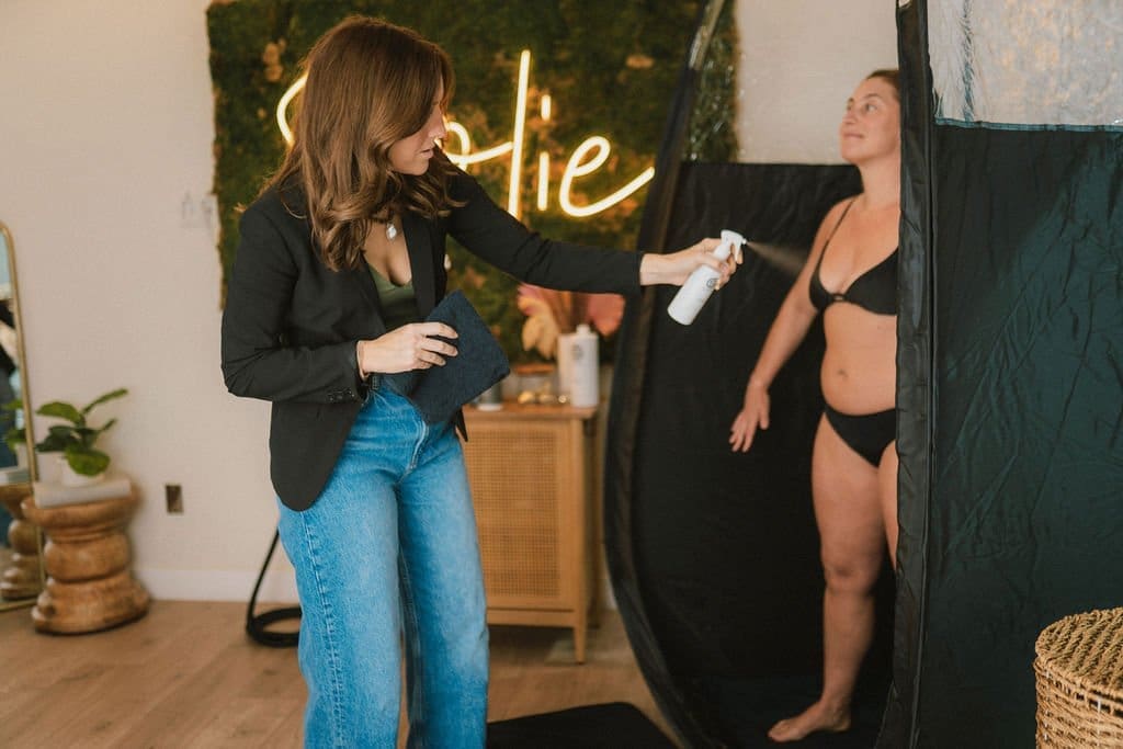 Spray tan technician applying pH Balancer Prep Spray to client in professional spray tan booth