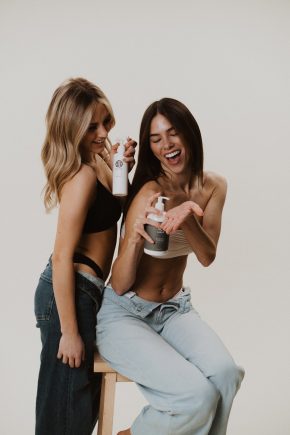 Two friends laughing while holding Sjolie pH Balancer spray and Barrier Cream pump bottle