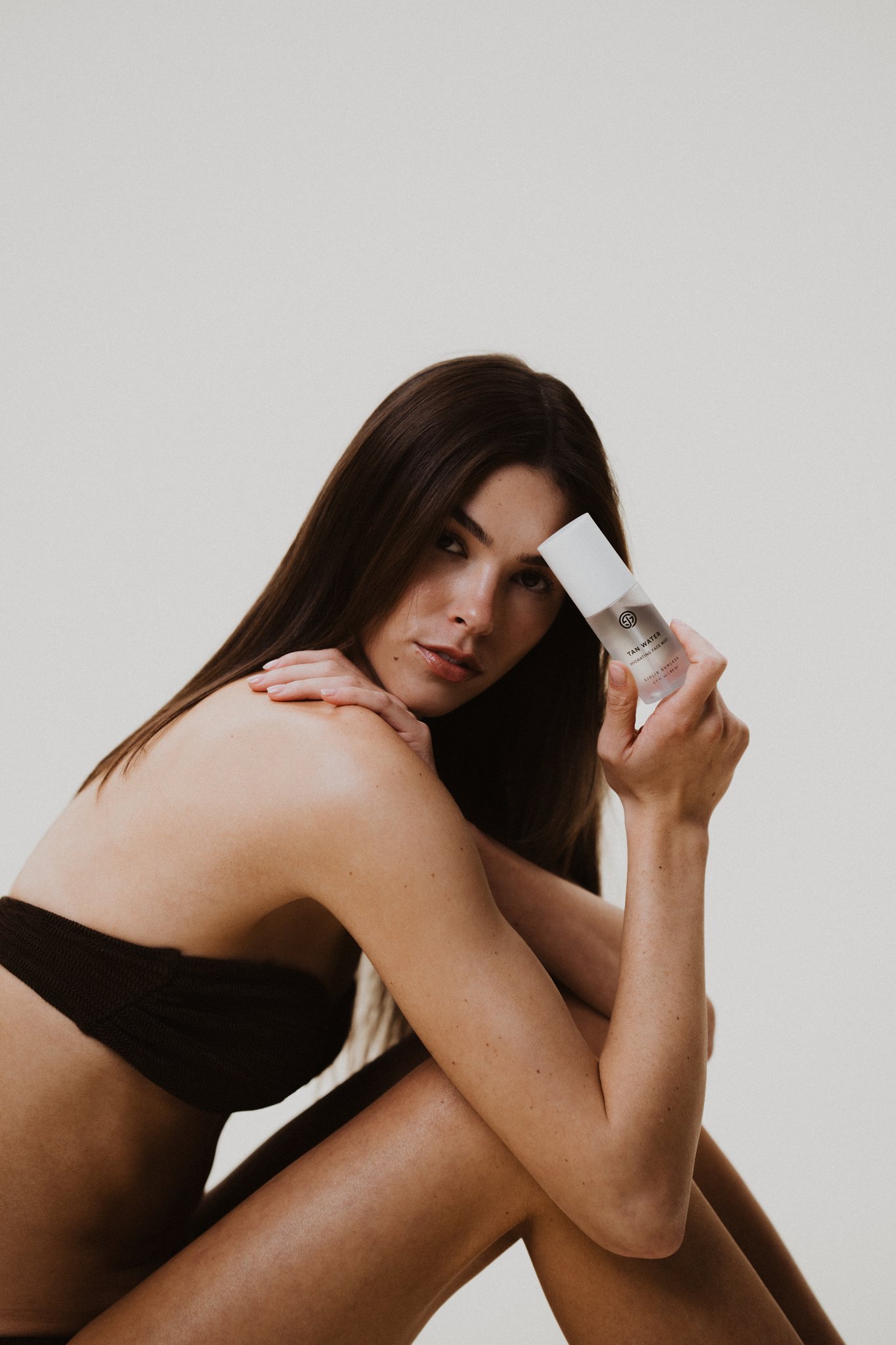 Woman resting her chin on her knee while holding a Sjolie Tan Water bottle near her face